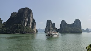 A scenic view of towering limestone islands covered with lush greenery rises from calm, turquoise waters. A traditional boat with a flag and several passengers sails across the water, creating a serene landscape.