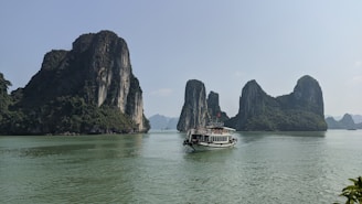 A scenic view of towering limestone islands covered with lush greenery rises from calm, turquoise waters. A traditional boat with a flag and several passengers sails across the water, creating a serene landscape.