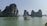 A scenic view of towering limestone islands covered with lush greenery rises from calm, turquoise waters. A traditional boat with a flag and several passengers sails across the water, creating a serene landscape.
