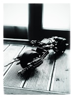 A small, scale model of a motorcycle made from plastic building blocks is positioned on a textured wooden surface. The lighting creates high contrast and shadows, emphasizing the lines and details of the model. The scene is backlit by a large window, providing a diffused light effect.