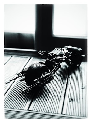 A small, scale model of a motorcycle made from plastic building blocks is positioned on a textured wooden surface. The lighting creates high contrast and shadows, emphasizing the lines and details of the model. The scene is backlit by a large window, providing a diffused light effect.