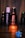 A bottle of Fogg perfume labeled 'Intense Oriental' is prominently displayed, backlit with a warm glow, creating a focus on its elegant red and black design. The background is dim, with other less distinct Fogg bottles visible, suggesting a collection or display. A blurred blue flower-like object appears in the foreground.