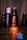 A bottle of Fogg perfume labeled 'Intense Oriental' is prominently displayed, backlit with a warm glow, creating a focus on its elegant red and black design. The background is dim, with other less distinct Fogg bottles visible, suggesting a collection or display. A blurred blue flower-like object appears in the foreground.