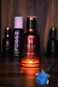 A bottle of Fogg perfume labeled 'Intense Oriental' is prominently displayed, backlit with a warm glow, creating a focus on its elegant red and black design. The background is dim, with other less distinct Fogg bottles visible, suggesting a collection or display. A blurred blue flower-like object appears in the foreground.