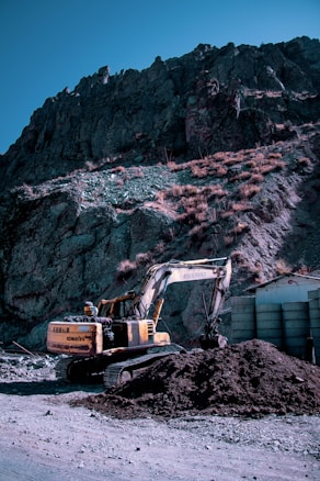 An excavator is parked on a dirt road at the base of a rocky mountain. It appears to be in the process of moving a pile of soil or rubble. The mountain is steep and rugged, with sparse vegetation. The sky is clear and bright, suggesting daylight.