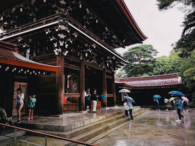 A traditional Japanese shrine with wooden architecture and paper decorations, surrounded by lush green trees. People are walking around the wet stone courtyard with umbrellas, suggesting a rainy day. The architectural details are ornate, reflecting cultural heritage.
