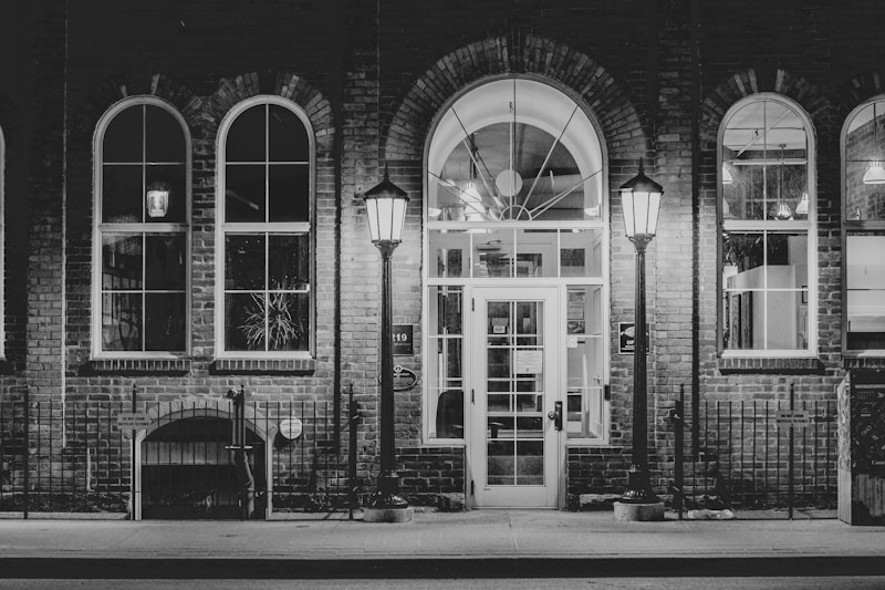 A nighttime scene features an elegant brick building facade with large windows and a central glass doorway. Two vintage-style street lamps illuminate the entrance, casting soft light. The setting is calm and the architecture highlights classic design elements.