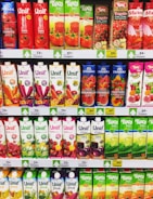 A supermarket shelf displaying a variety of juice cartons. The top row features red and green cartons with tomato and mixed fruit juices. Below are rows of multicolored cartons with flavors including watermelon, vegetable, and pomegranate. Each row is labeled with prices in yellow and white tags.