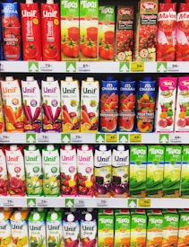A supermarket shelf displaying a variety of juice cartons. The top row features red and green cartons with tomato and mixed fruit juices. Below are rows of multicolored cartons with flavors including watermelon, vegetable, and pomegranate. Each row is labeled with prices in yellow and white tags.