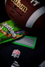 A close-up shot of a vintage brown leather American football with visible white laces and branding, partially covering a colorful Skittles candy packet. Below these items, part of a dark colored NFL jersey with a prominent NFL logo patch is visible.