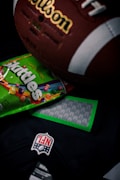 A close-up shot of a vintage brown leather American football with visible white laces and branding, partially covering a colorful Skittles candy packet. Below these items, part of a dark colored NFL jersey with a prominent NFL logo patch is visible.
