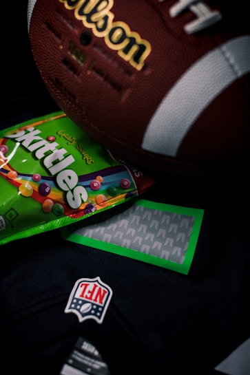 A close-up shot of a vintage brown leather American football with visible white laces and branding, partially covering a colorful Skittles candy packet. Below these items, part of a dark colored NFL jersey with a prominent NFL logo patch is visible.