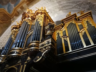A grand and ornate pipe organ with intricate gold detailing and large vertical pipes against a decorated ceiling. The organ displays elaborate carvings and artistic craftsmanship, contributing to its majestic and imposing presence.