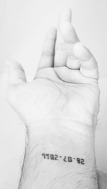 A human hand is prominently positioned, palm facing up, with fingers slightly curled. The wrist displays a minimalist tattoo featuring a date printed in a bold, sans-serif font. The image is in black and white, accentuating the contrast between the skin and the tattoo.