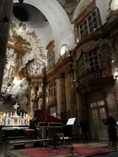 A richly decorated baroque interior with ornate sculptures and elaborate architectural details. Gold and marble elements accentuate the grandeur of the space. A musician is playing a harpsichord, and a music stand with sheet music is visible. The setting has a large altar with candles and a religious motif.
