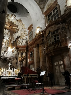 A richly decorated baroque interior with ornate sculptures and elaborate architectural details. Gold and marble elements accentuate the grandeur of the space. A musician is playing a harpsichord, and a music stand with sheet music is visible. The setting has a large altar with candles and a religious motif.