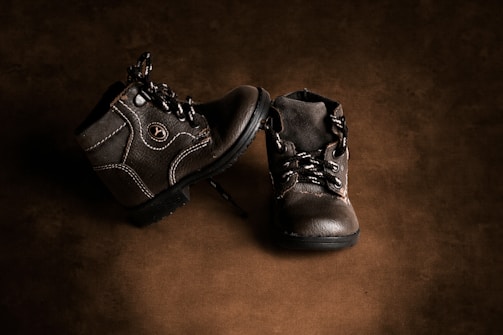 A pair of small, worn leather boots with detailed stitching and laces are arranged on a textured brown background. The boots have a rugged appearance and feature a visible logo on the side.