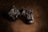 A pair of small, worn leather boots with detailed stitching and laces are arranged on a textured brown background. The boots have a rugged appearance and feature a visible logo on the side.
