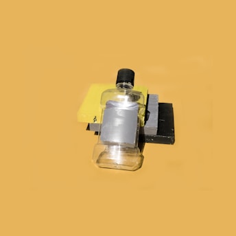 A clear plastic bottle with a black cap is lying on its side on top of stacked rectangular objects in shades of yellow, gray, and black. The composition casts distinct shadows and is set against a plain, solid background.