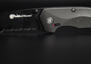 A close-up view of a serrated, black-colored knife with the brand Smith & Wesson engraved on its blade. The handle is metallic grey, featuring a circular emblem and a small red dot near a textured grip area.