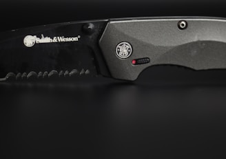 A close-up view of a serrated, black-colored knife with the brand Smith & Wesson engraved on its blade. The handle is metallic grey, featuring a circular emblem and a small red dot near a textured grip area.