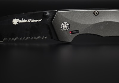 A close-up view of a serrated, black-colored knife with the brand Smith & Wesson engraved on its blade. The handle is metallic grey, featuring a circular emblem and a small red dot near a textured grip area.