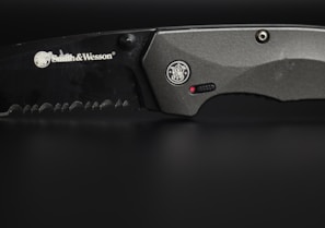 A close-up view of a serrated, black-colored knife with the brand Smith & Wesson engraved on its blade. The handle is metallic grey, featuring a circular emblem and a small red dot near a textured grip area.