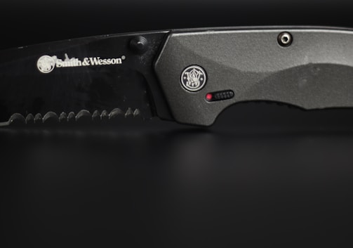 A close-up view of a serrated, black-colored knife with the brand Smith & Wesson engraved on its blade. The handle is metallic grey, featuring a circular emblem and a small red dot near a textured grip area.