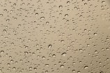Water droplets of varying sizes scattered across a smooth glass or plastic surface, creating a pattern against a neutral beige or light brown background.