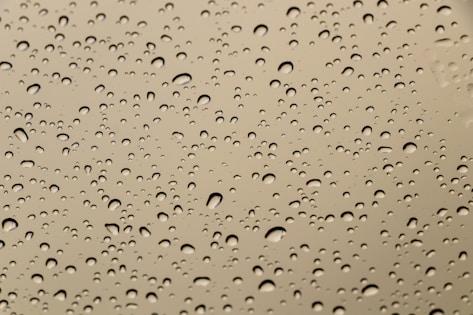 Water droplets of varying sizes scattered across a smooth glass or plastic surface, creating a pattern against a neutral beige or light brown background.