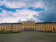 A grand, historic palace with a yellow facade and lined with rows of evenly spaced windows and decorative statues on the roof. The foreground features an expansive courtyard with neatly trimmed gardens. Overhead, the sky is expansive, with a mix of bright blue and large, dramatic clouds.