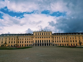 A grand, historic palace with a yellow facade and lined with rows of evenly spaced windows and decorative statues on the roof. The foreground features an expansive courtyard with neatly trimmed gardens. Overhead, the sky is expansive, with a mix of bright blue and large, dramatic clouds.