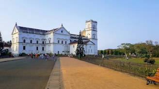 A historic, white church building with classical architecture, featuring arched windows and a tall bell tower. The foreground includes a paved pathway and benches, while several people are walking around the area. The background shows well-maintained lawns and trees under a clear, blue sky.