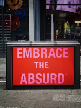 A street scene featuring a brightly colored red sign with bold pink letters reading 'EMBRACE THE ABSURD'. The sign is set against the window of a building. Behind the glass are abstract designs and gears, along with visible mannequins displaying clothing.