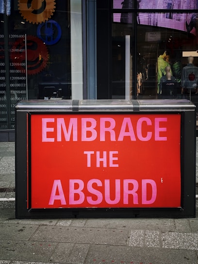A street scene featuring a brightly colored red sign with bold pink letters reading 'EMBRACE THE ABSURD'. The sign is set against the window of a building. Behind the glass are abstract designs and gears, along with visible mannequins displaying clothing.