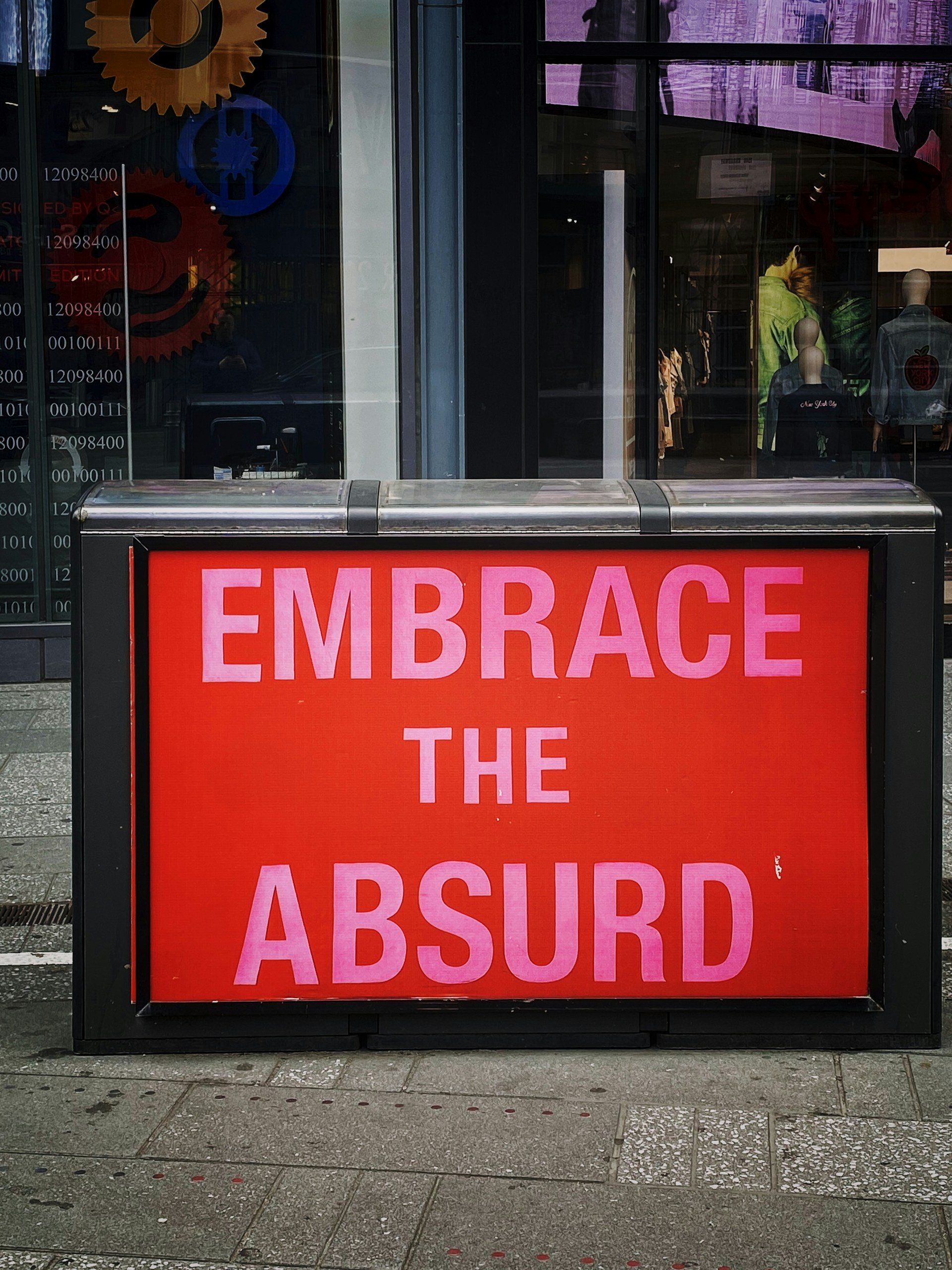 A street scene featuring a brightly colored red sign with bold pink letters reading 'EMBRACE THE ABSURD'. The sign is set against the window of a building. Behind the glass are abstract designs and gears, along with visible mannequins displaying clothing.