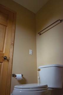 A small, clean bathroom corner with beige walls. A white toilet with a closed lid is positioned near the center. Above the toilet is an empty towel rack attached to the wall. Next to the toilet is a wooden door with a silver handle, and a roll of toilet paper hangs on a wall-mounted holder.