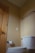 A small, clean bathroom corner with beige walls. A white toilet with a closed lid is positioned near the center. Above the toilet is an empty towel rack attached to the wall. Next to the toilet is a wooden door with a silver handle, and a roll of toilet paper hangs on a wall-mounted holder.