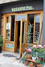 A small storefront with a sign reading 'Key Lime Pie' in green, wooden letters. The shop has wooden framed windows and doors, with one door open, inviting entry. There are various potted plants and decorative flamingo statues outside. A mosaic of a wave is featured near the lower part of the window. A sign offers cold brew iced coffee, and the atmosphere appears quaint and welcoming.