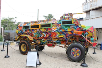 A heavily modified, armored vehicle covered in colorful geometric patterns stands on display. The artwork features vibrant and intricate designs in a wide array of colors, including reds, blues, yellows, and greens. Surrounding the vehicle are black stanchions and a display plaque, indicating its status as an exhibit. In the background, there are trees, a fence, and shipping containers, suggesting an outdoor setting.