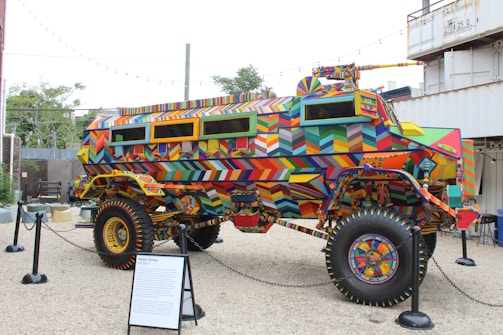 A heavily modified, armored vehicle covered in colorful geometric patterns stands on display. The artwork features vibrant and intricate designs in a wide array of colors, including reds, blues, yellows, and greens. Surrounding the vehicle are black stanchions and a display plaque, indicating its status as an exhibit. In the background, there are trees, a fence, and shipping containers, suggesting an outdoor setting.