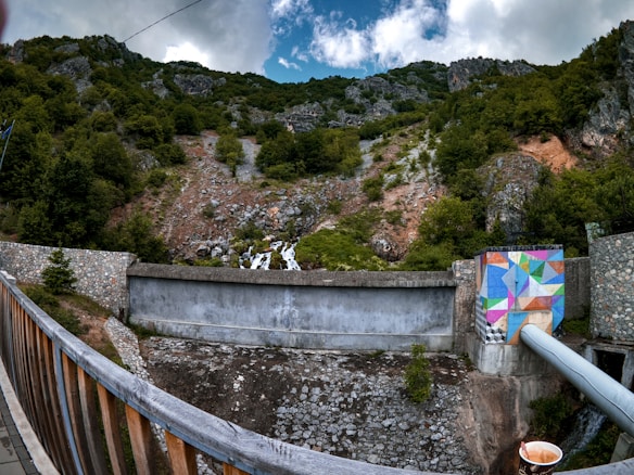 A scenic view features a stone and concrete dam or retaining wall against a backdrop of a forested mountain. The vegetation is dense with green trees and shrubs interspersed with rocky outcrops. A brightly colored mural with geometric shapes decorates part of the structure, adding a splash of vibrant color, and a metallic pipe extends from the painted section. The foreground includes a wooden railing, and a disposable cup with a drink is visible on the right.