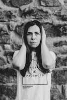 A woman stands against a stone wall, holding her hands to the sides of her head. She has a serious expression on her face. Her t-shirt displays the text 'People ≠ Products' and '#StopTheDemand'. The image is in black and white, adding a dramatic effect.