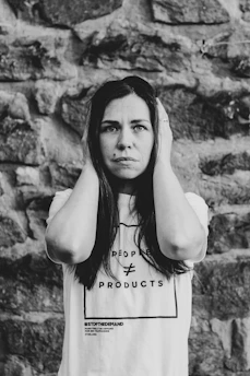 A woman stands against a stone wall, holding her hands to the sides of her head. She has a serious expression on her face. Her t-shirt displays the text 'People ≠ Products' and '#StopTheDemand'. The image is in black and white, adding a dramatic effect.