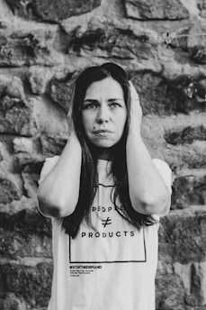 A woman stands against a stone wall, holding her hands to the sides of her head. She has a serious expression on her face. Her t-shirt displays the text 'People ≠ Products' and '#StopTheDemand'. The image is in black and white, adding a dramatic effect.