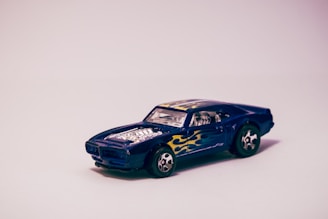 A toy car modeled after a classic muscle car with a dark blue body featuring yellow flame decals on the sides. The car has chrome-like details on the hood, resembling an engine, along with shiny wheels that have a star-like design. The background is a soft gradient from pink to white, providing a clean and focused backdrop.