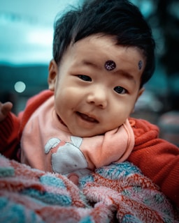 A smiling baby with black hair gazes warmly at the camera, wearing a pink bib with a rabbit design and a red jacket. The baby is wrapped in a colorful, textured blanket.