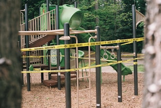 A children's playground featuring a green slide, climbing ladder, and gymnastic rings is cordoned off with yellow caution tape. The area is surrounded by trees, suggesting a park setting.