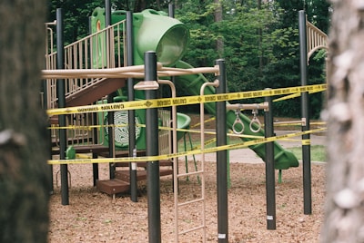 A children's playground featuring a green slide, climbing ladder, and gymnastic rings is cordoned off with yellow caution tape. The area is surrounded by trees, suggesting a park setting.