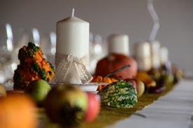 A decorative table arrangement featuring a row of white pillar candles, surrounded by various small gourds and fruits. The candles are adorned with lace-ribbon, and soft lighting creates a warm, inviting atmosphere.